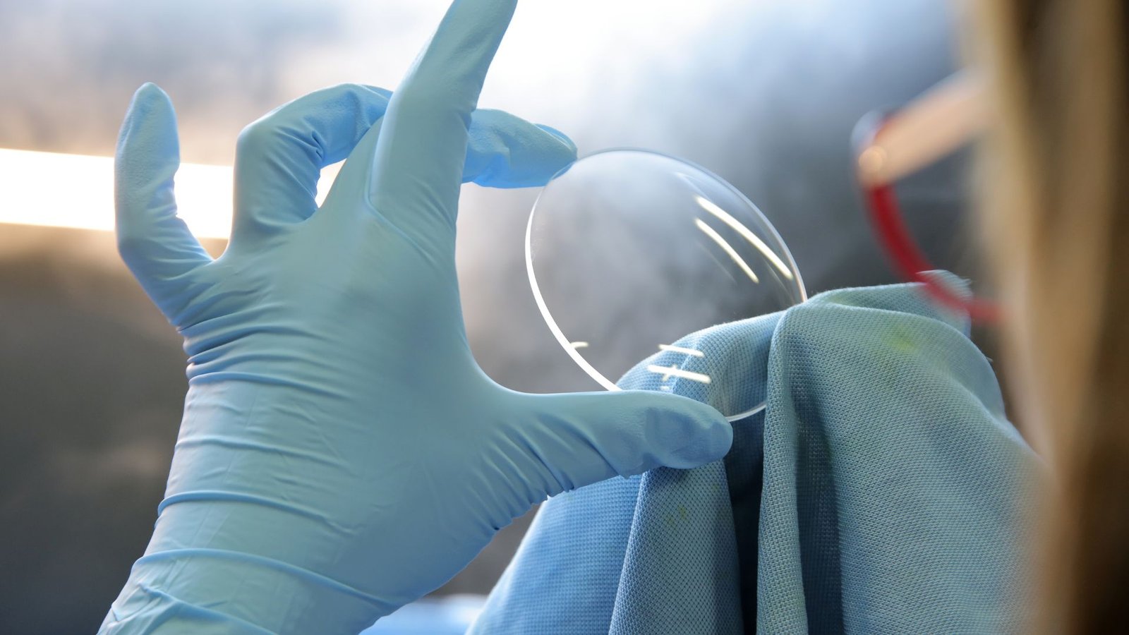 Main gantée tenant un verre ophtalmique devant une lumière, dans l’atelier Persepolis en Guadeloupe